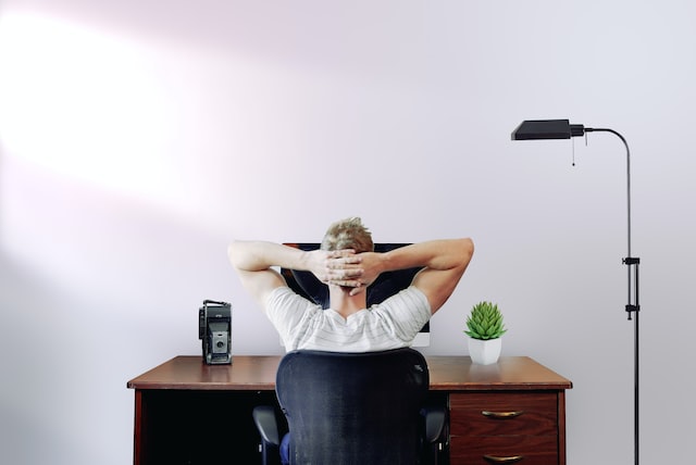 homme de dos assis à son bureau face à son ordinateur, les bras croisés derrière la tête