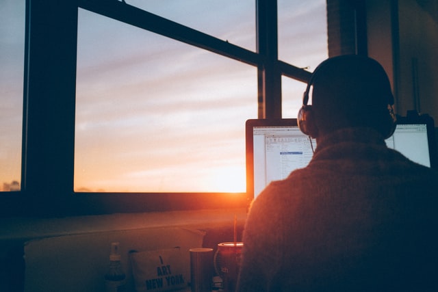 homme de dos face à un écran d'ordinateur, casque sur les oreille, derrière une fenêtre au travers de laquelle luit un coucher de soleil